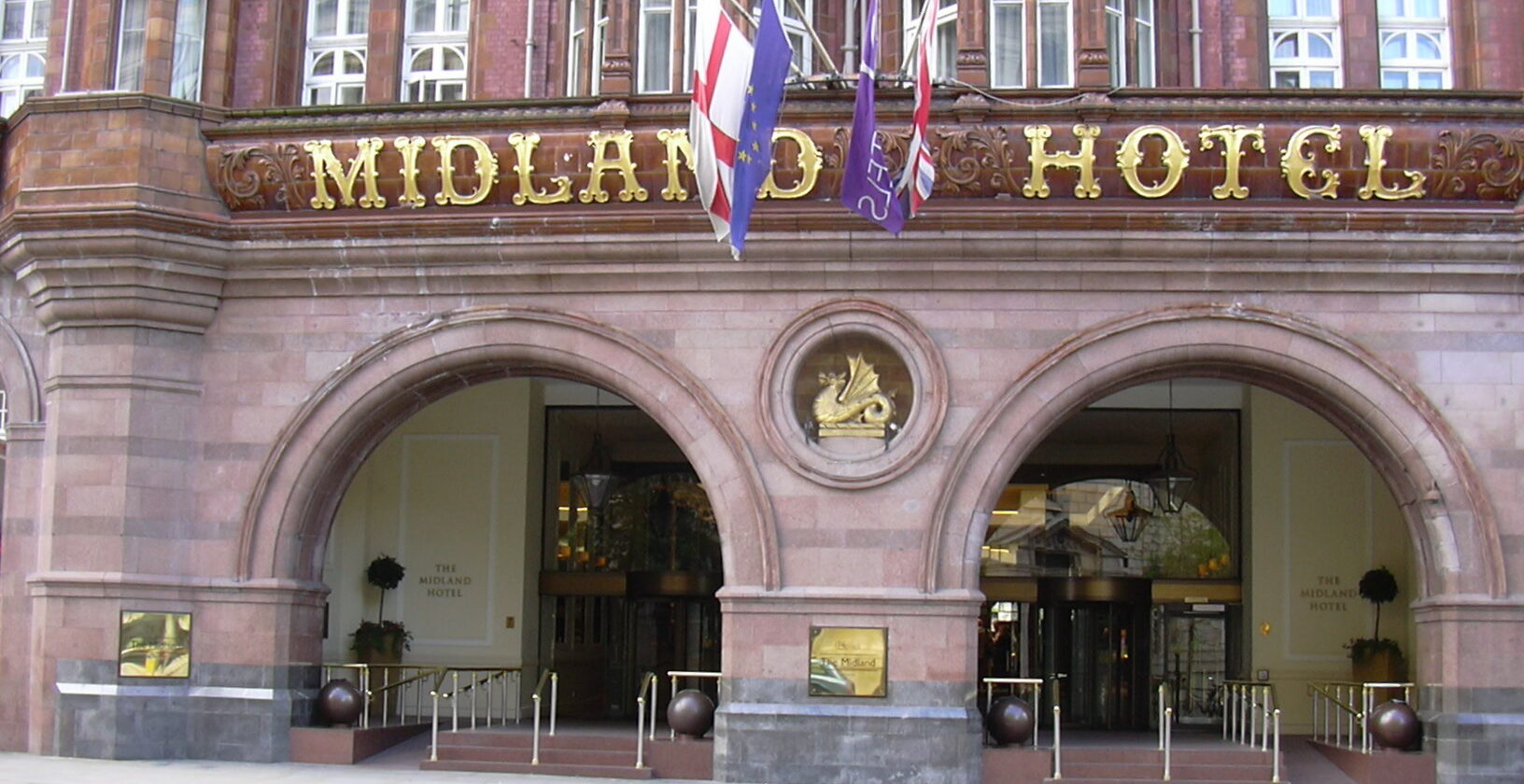 The arched stone entrance of the Midland Hotel on Peter Street in Manchester, with gold MIDLAND HOTEL lettering above the doors and flags on the facade