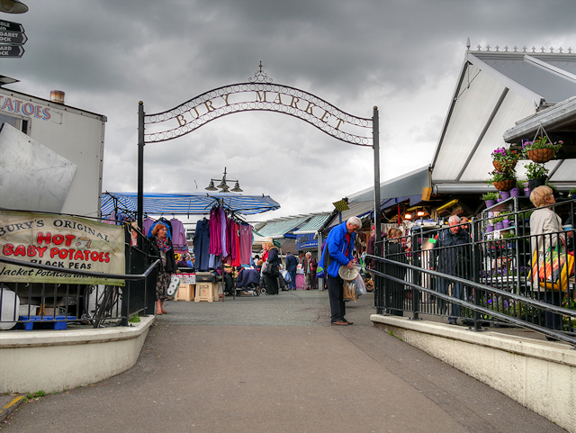The iron Bury Market arch over the entrance, with a hot baby potatoes and black peas stall and shoppers beyond
