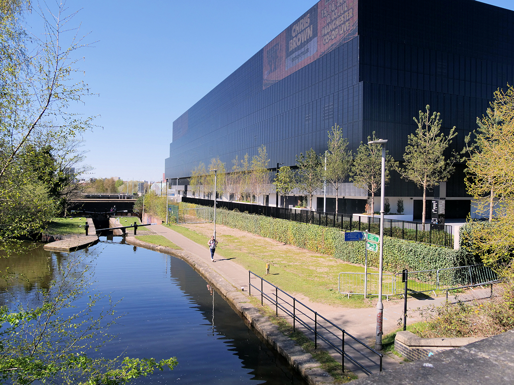 The black-clad exterior of the Co-op Live arena seen across the Ashton Canal on a clear day, with a event banner on the upper facade and a towpath in the foreground