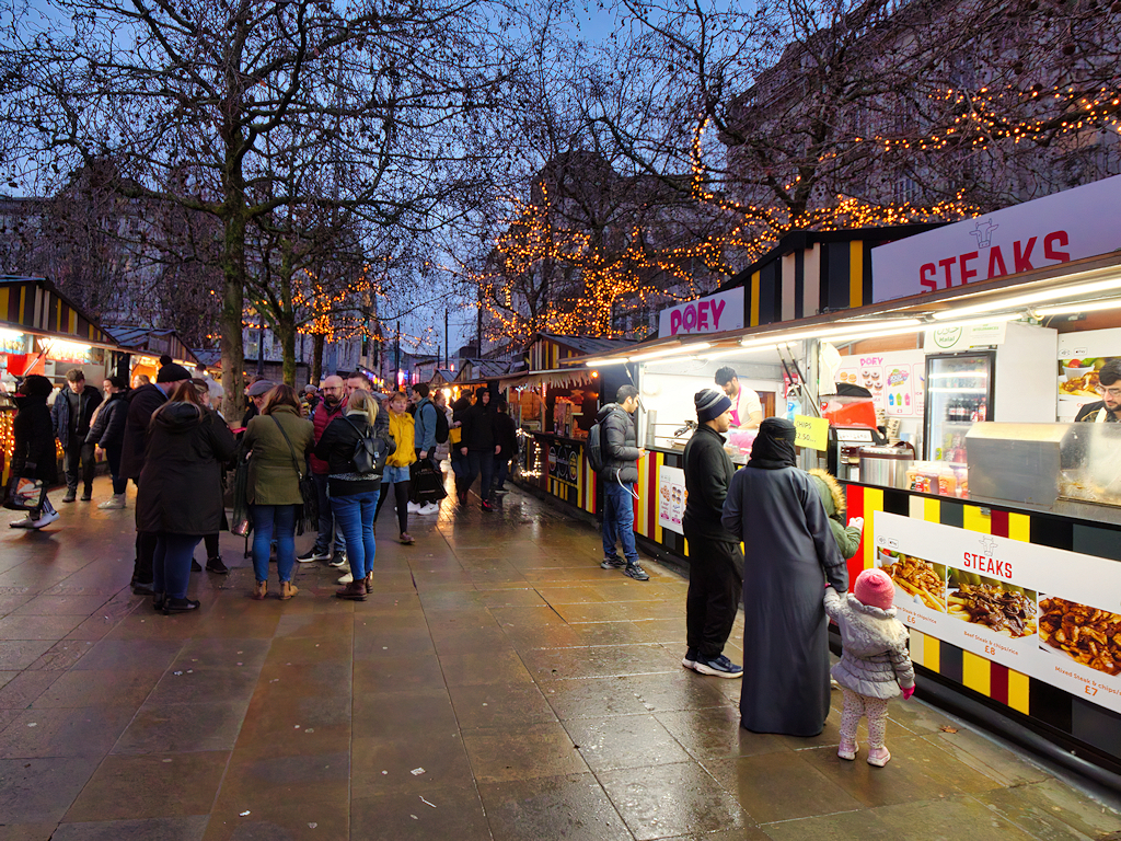 Manchester Christmas Markets Food & Drink: What to Eat & Drink 2 Families queuing at hot food stalls along the paths of Piccadilly Gardens Christmas market