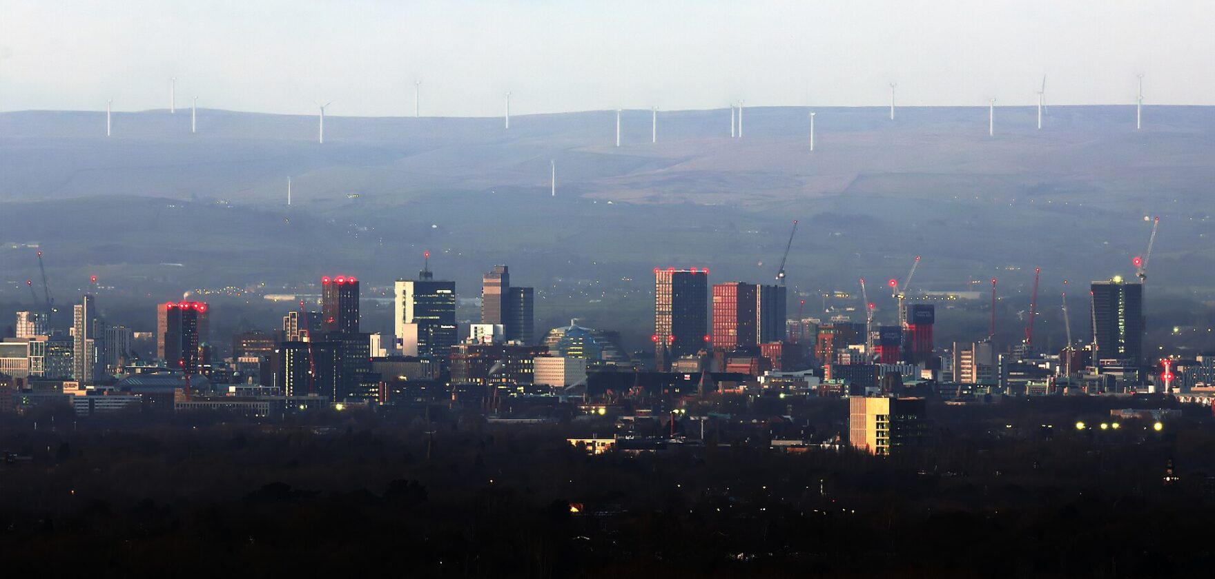 Manchester city-centre skyline at dusk in late February with the Pennine moors and wind turbines behind