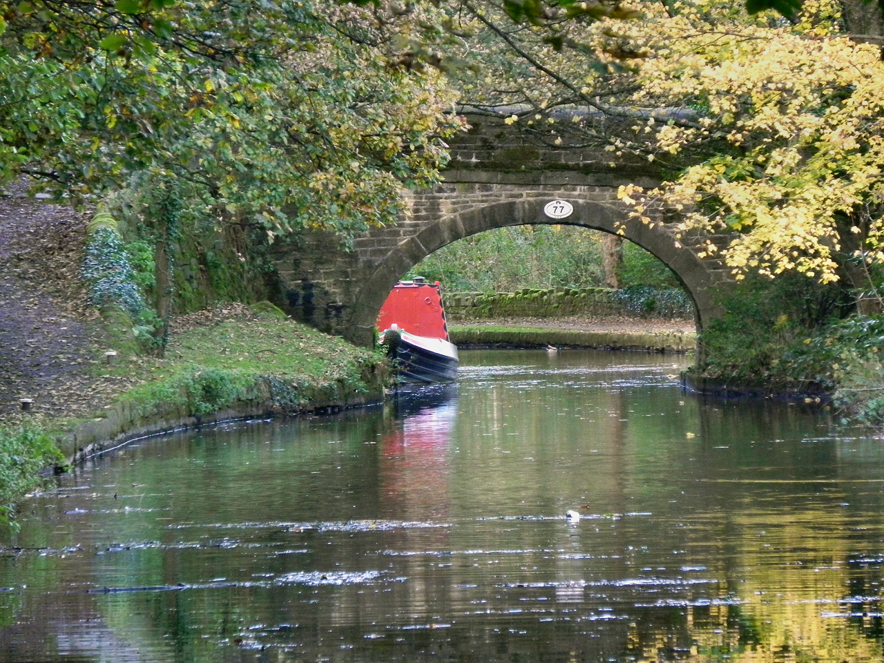 A red narrowboat approaching a stone bridge on the Huddersfield Narrow Canal in autumn