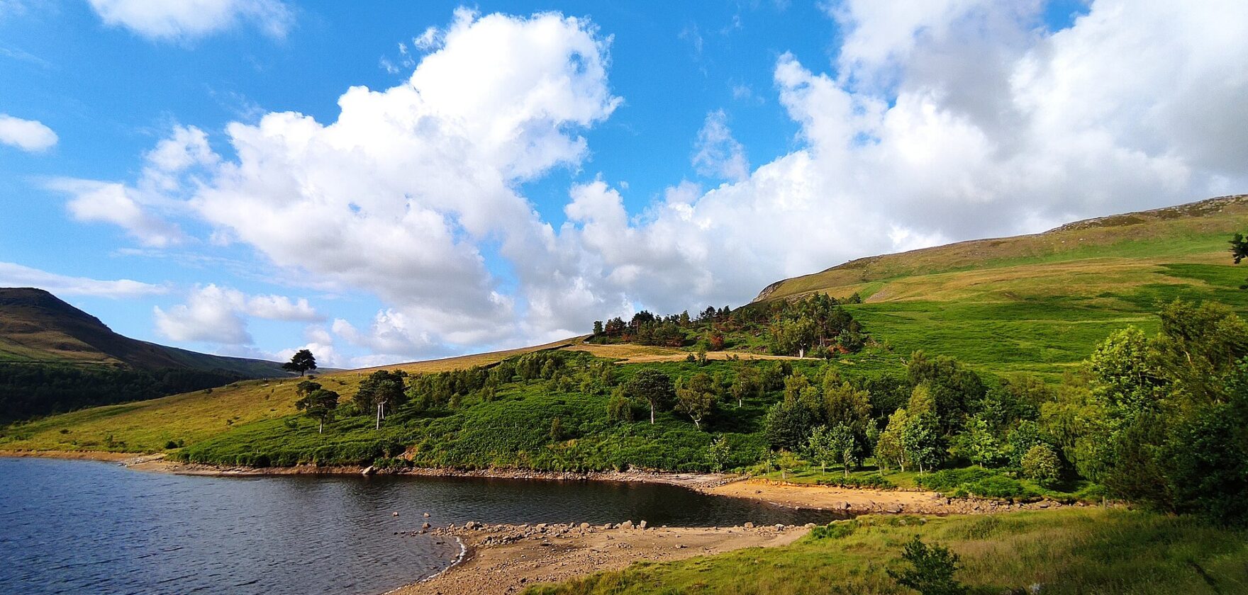 Dovestone Reservoir shoreline beneath green moorland slopes and a blue summer sky