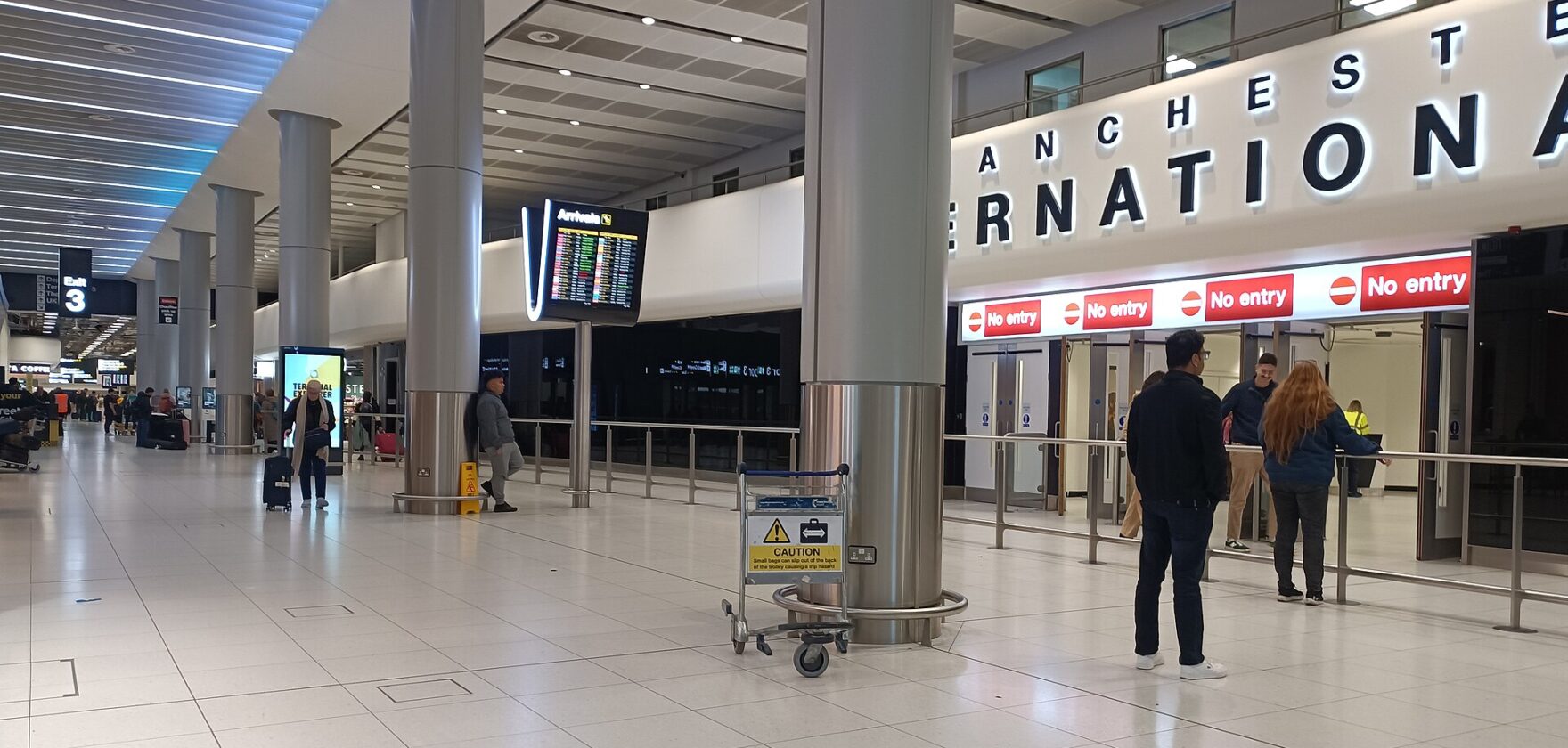 The Terminal 2 arrivals hall at Manchester Airport with a large Manchester International sign, exit doors and passengers with trolleys