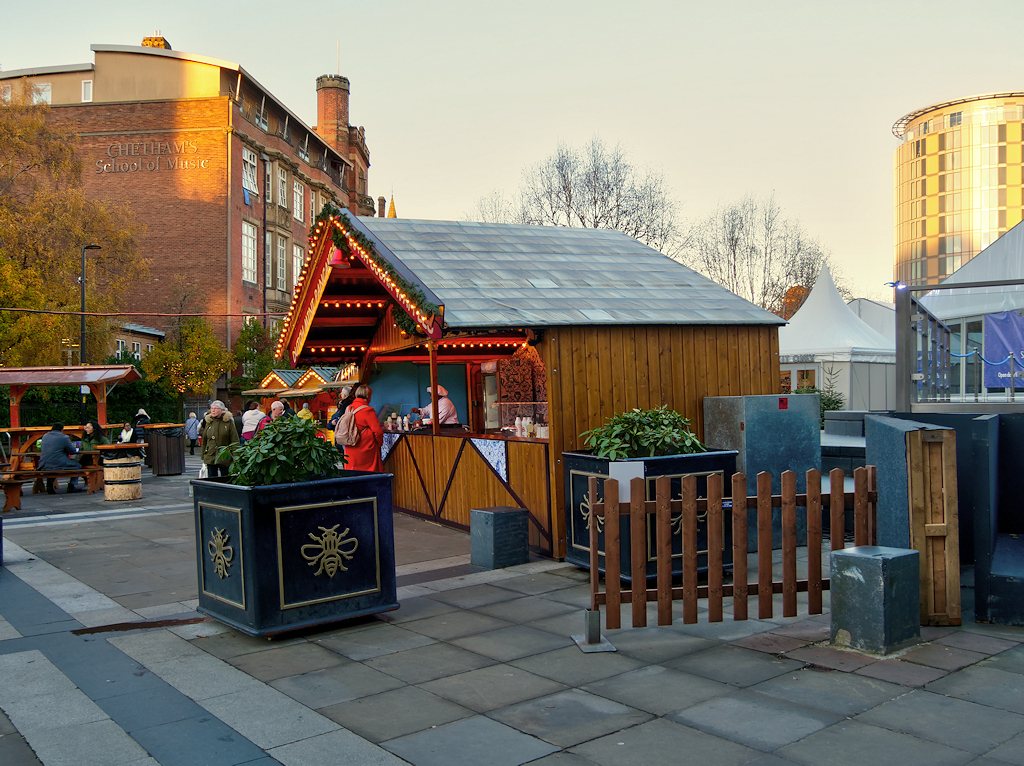 Wooden Christmas market chalets behind a picket fence and a planter decorated with the Manchester bee in Cathedral Gardens