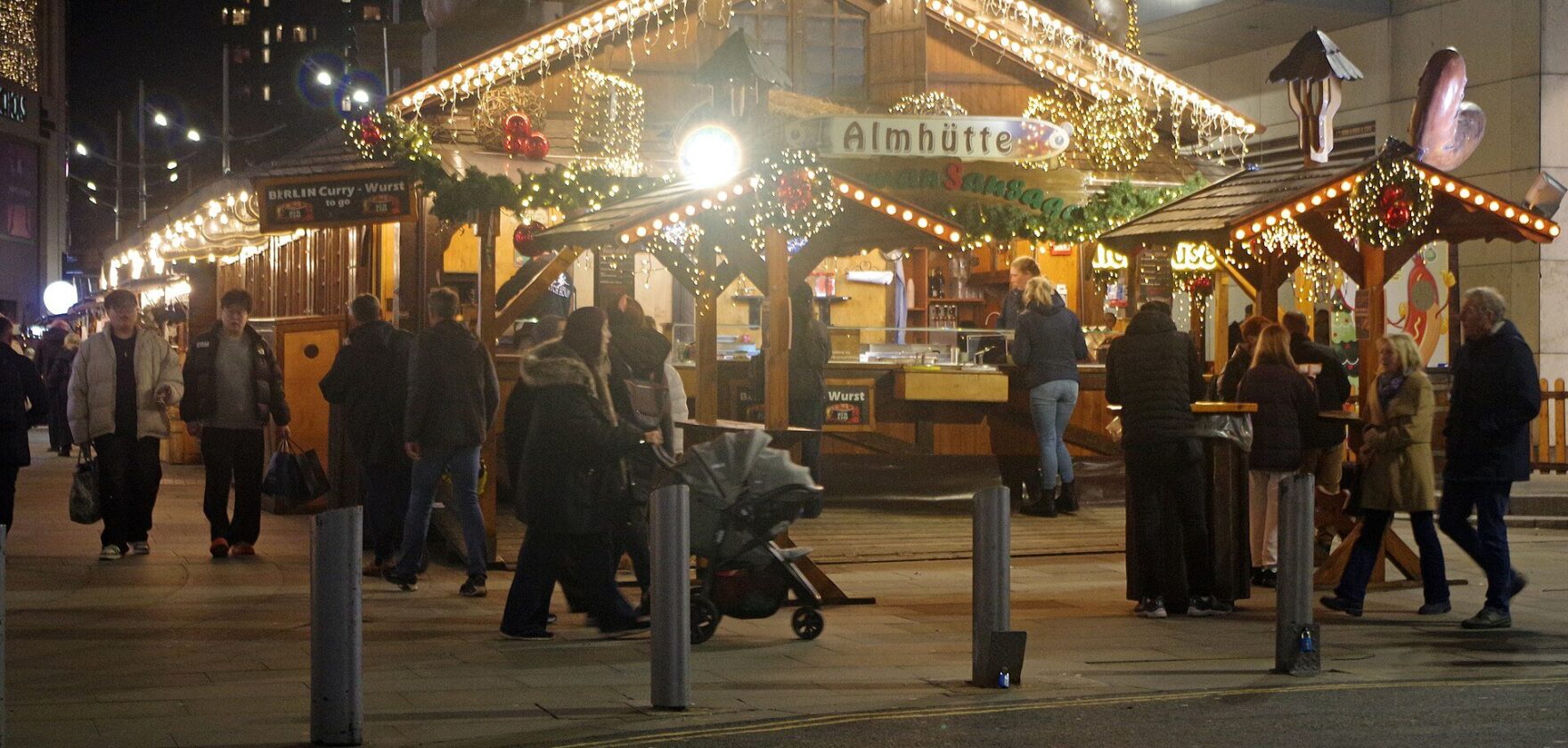 Wooden market chalets strung with fairy lights and shoppers walking past at the Manchester Christmas Markets at night